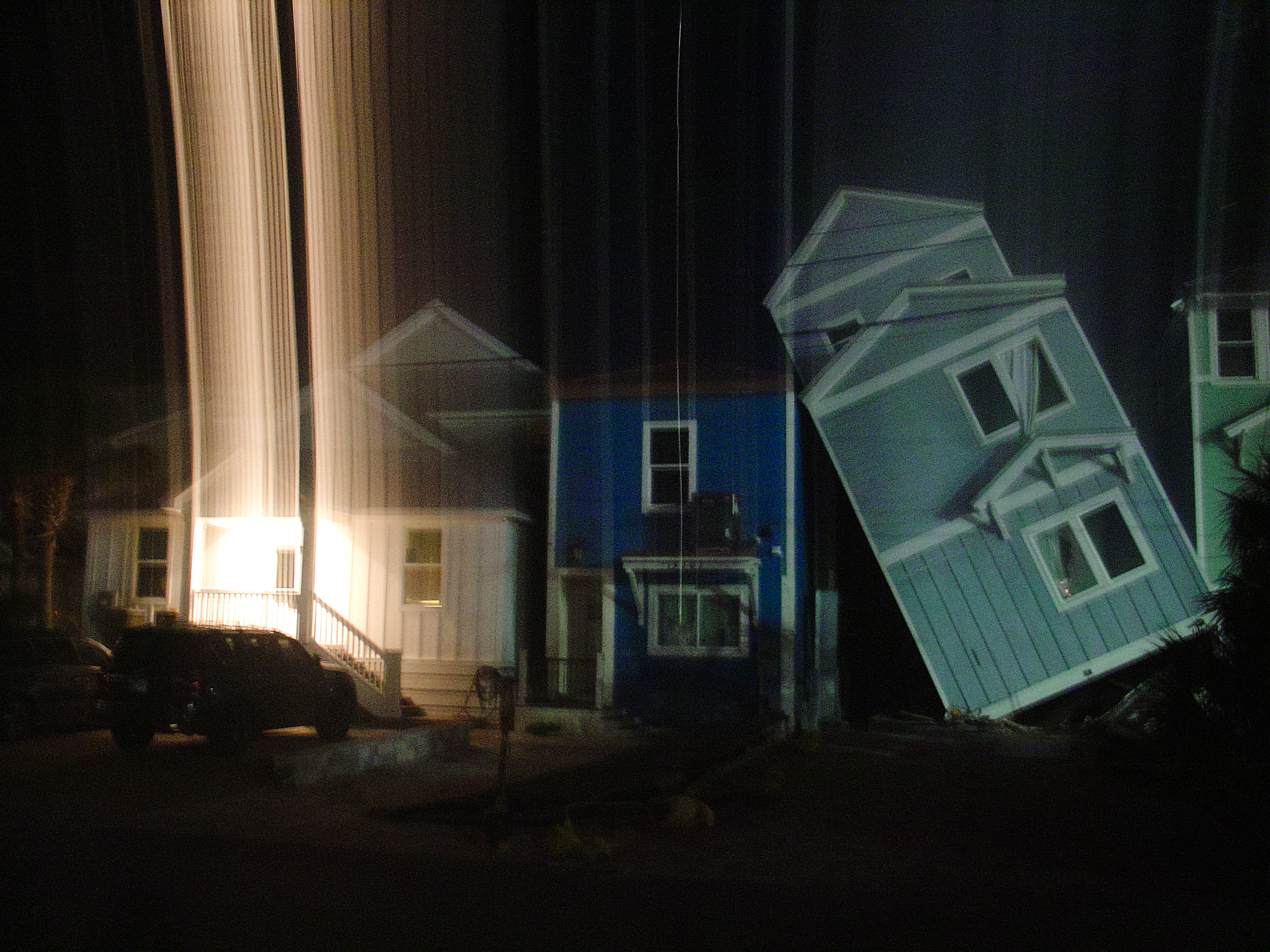 A row of houses at night, vertical light streaks from a long exposure smearing one pale house sideways, a dark SUV parked at the curb