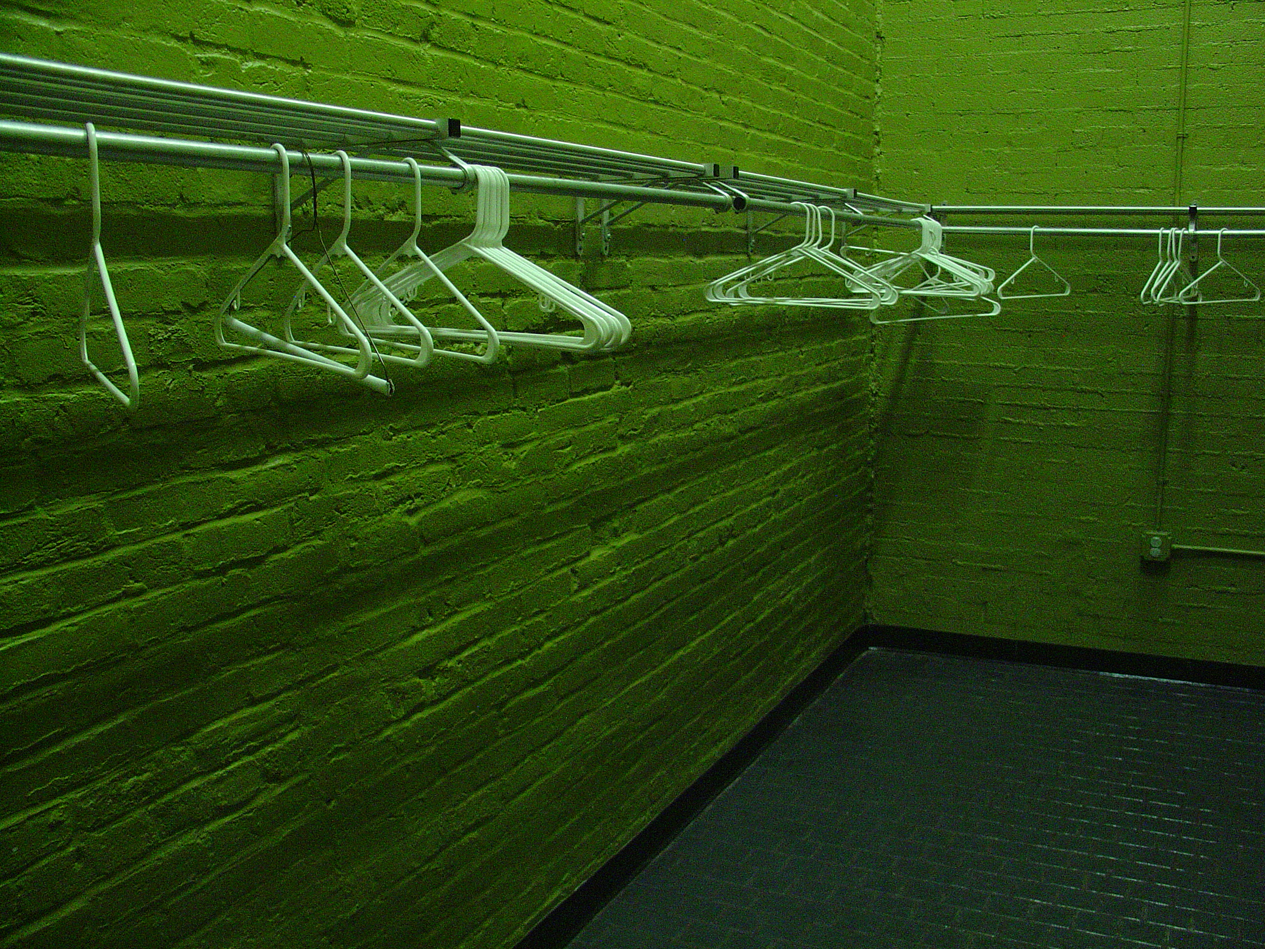 An empty coat-check rail of white wire hangers along a brick wall washed entirely green by overhead light