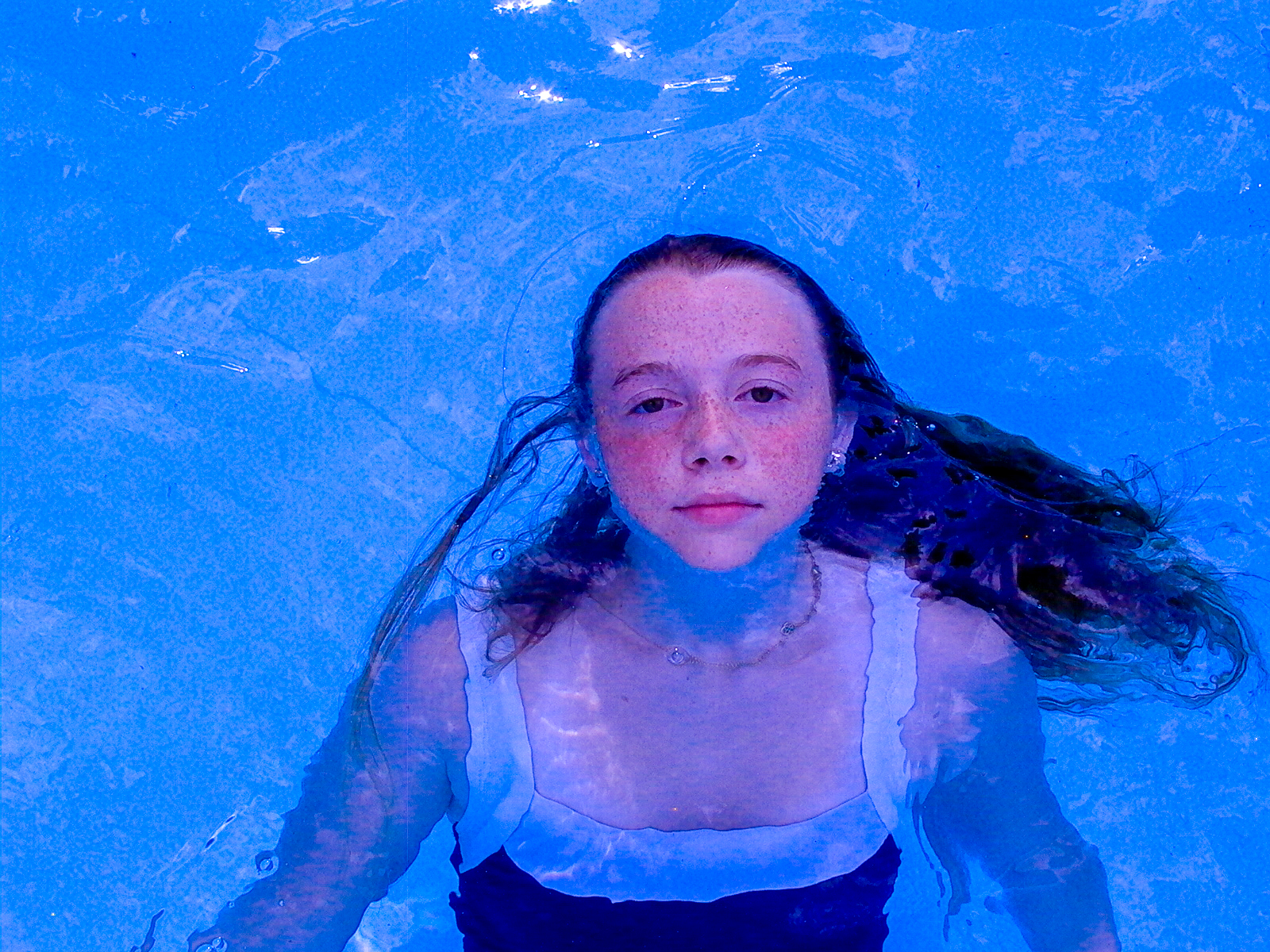 A girl submerged to the shoulders in vivid blue pool water, hair fanning out around her, looking up into the camera