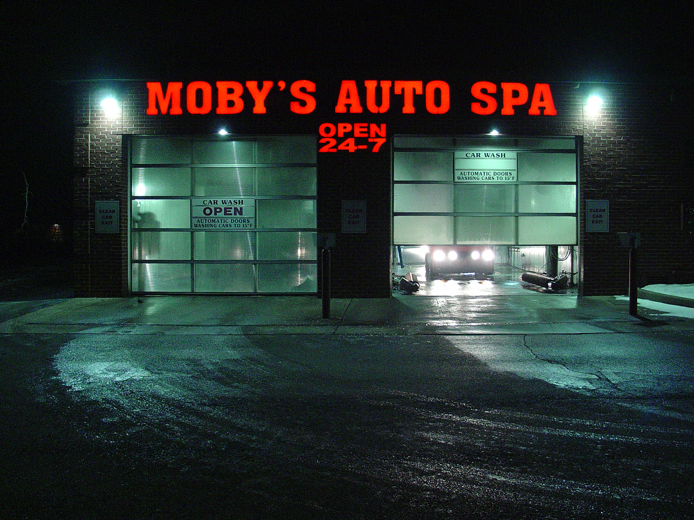 A car wash at night, red letters and green-blue glass lighting the wet pavement