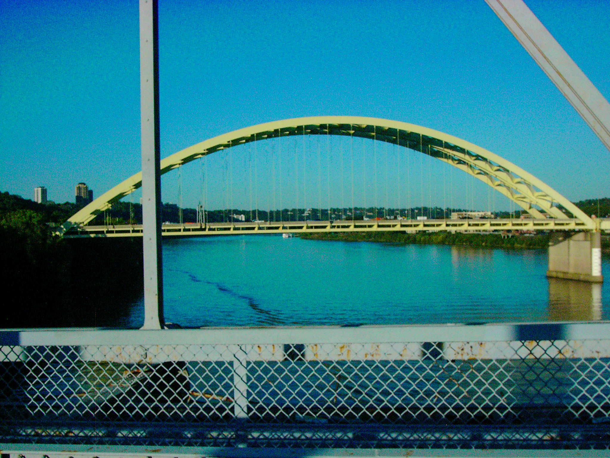 A pale arch bridge seen through a gridded window or fence, river and blue-green foreground split by hard lines