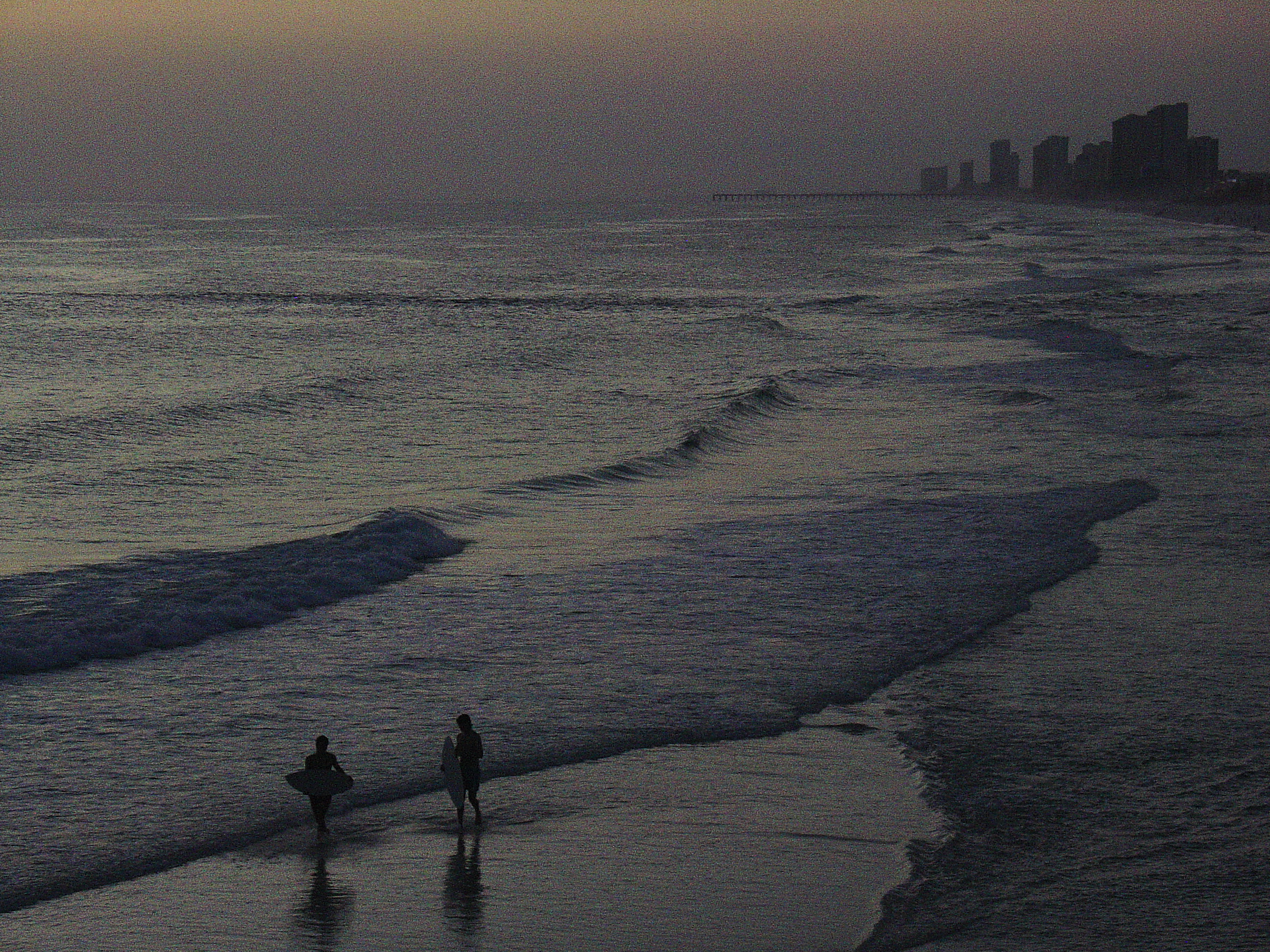 Two figures carrying boards at the waterline in low light, a pier and high-rise towers dissolving into haze down the shore, the sky a field of trembling grain